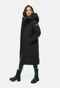 Black puffer coat with a long design, zip closure, large hood, and quilted texture. Paired with dark jeans and black ankle boots.