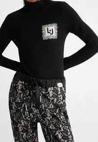 Pull à col roulé noir en tissu à motif de dentelle avec un écusson logo bordé de dentelle blanche, associé à un pantalon en dentelle noir avec cordon de serrage.