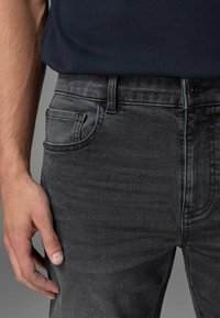 Graue Denim-Jeans mit schlankem Schnitt, ausgestattet mit einer einzelnen Gesäßtasche, Knopfverschluss und subtilen Verblassungen, kombiniert mit einem navyfarbenen Oberteil.
