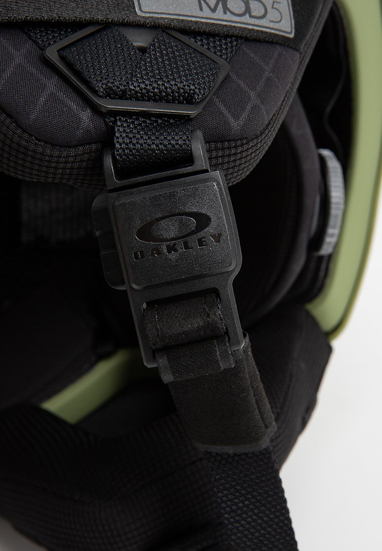 Correa de casco negra con una hebilla ajustable, que presenta un logo de Oakley. La correa tiene textura y está unida a una carcasa de casco verde.