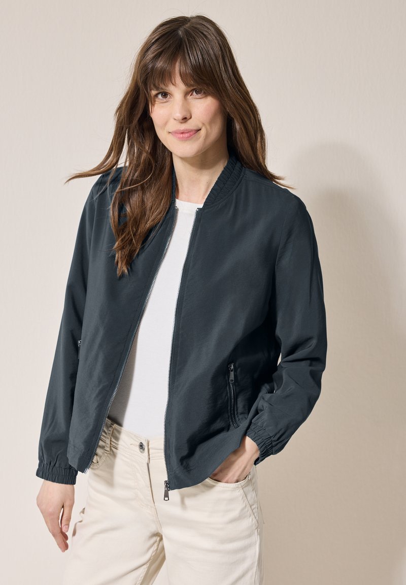 Marinblå bomberjacka i lätt tyg, med dragkedja fram, elastiska ärmslut och sidofickor; bärs över en vit topp.