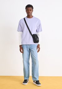 Lyse purpur t-shirt lagd over en hvid langærmet bluse, mellemblå jeans, sort crossbody taske og sorte sneakers med hvide accenter.
