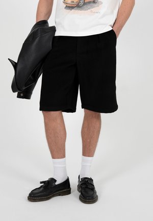 Hombre con pantalones cortos negros, calcetines blancos, mocasines negros con borlas, camiseta blanca con un estampado de coche, sosteniendo una chaqueta de cuero negra en una mano.