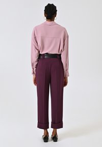 Blouse à manches longues rose clair avec un col, associée à un pantalon taille haute violet foncé avec des ourlets retroussés et une ceinture noire. Chaussures noires.