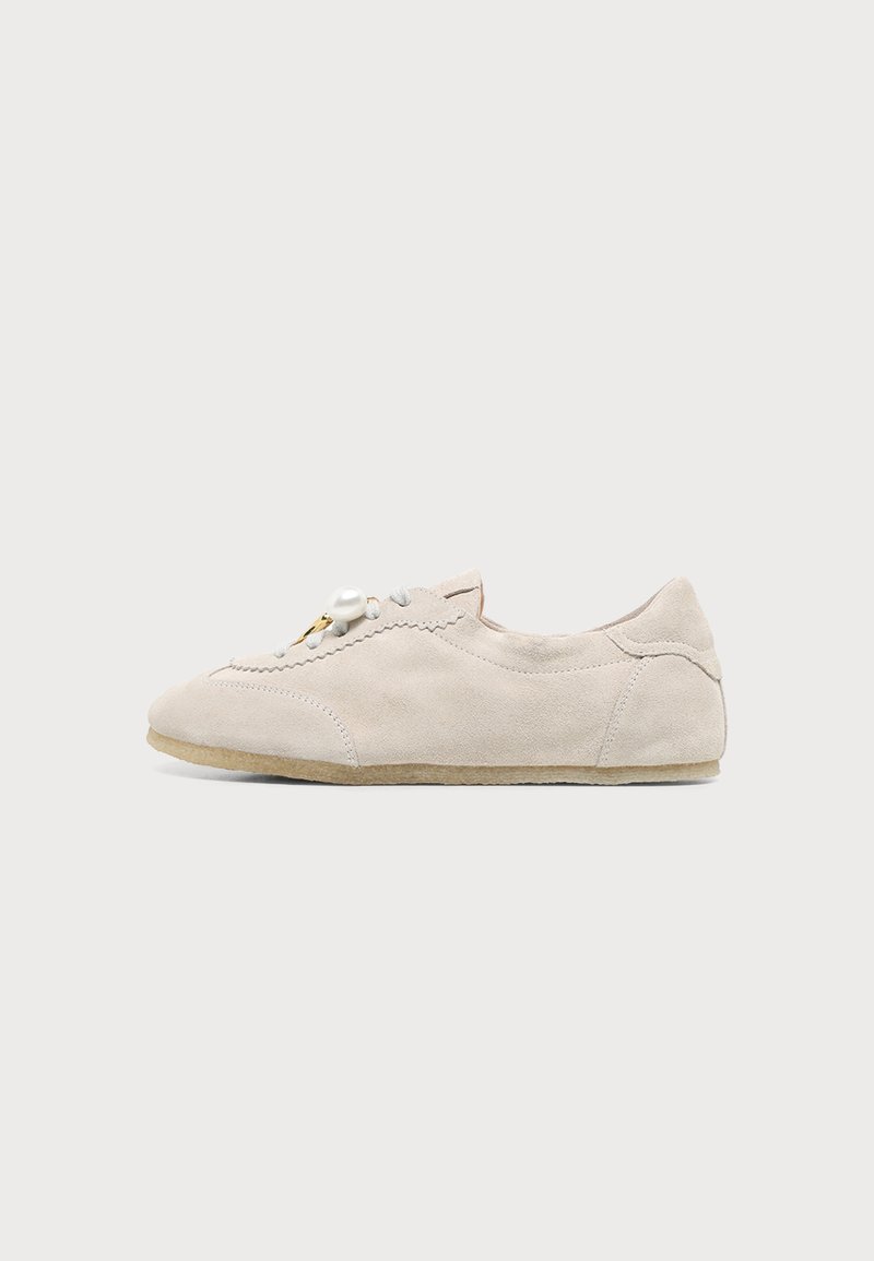 Chaussure plate en daim beige avec bord festonné, lacets à l’avant et grosse perle décorative sur fond gris.