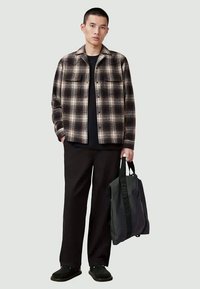 Karierte Flanellhemd in Braun und Creme, schwarzes T-Shirt, schwarze Hose, trägt eine schwarze Tragetasche. Einfaches Design mit zwei Brusttaschen.