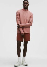 Model masculin îmbrăcat cu un pulover lung cu mânecă și fermoar la jumătate de culoare roz prăfuit, pantaloni scurți de culoare rugini, șosete albe până la gleznă și adidași albi, stând pe un fundal simplu.