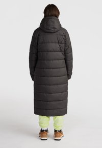 Lång svart puffjacka med huva, med en kviltad design och förlängd form. Modellen bär neongröna byxor och bruna skor.