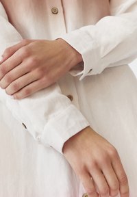 Bílá lněná košile s viditelnými knoflíky, ukazujícími vyhrnuté rukávy a složené manžety. Hladká textura s jemnými osvětlenými vzory.