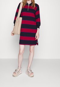Abito a righe in blu navy e rosso, con colletto con zip e maniche a tre quarti. Abbinato a una borsa a tracolla beige e sandali spessi.