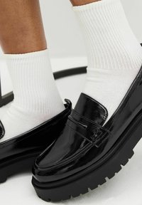 Zwarte lakleren loafers met een dikke, gestructureerde zool, gecombineerd met geribde witte enkelsokken. Glad oppervlak en slank ontwerp.