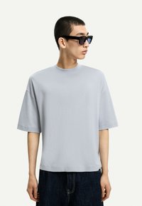 Svetlo siva majica s kratkimi rokavi in okroglim izrezom, izdelana iz gladkega materiala, v kombinaciji s temnimi kavbojkami. Črne sončna očala dopolnjujejo videz.