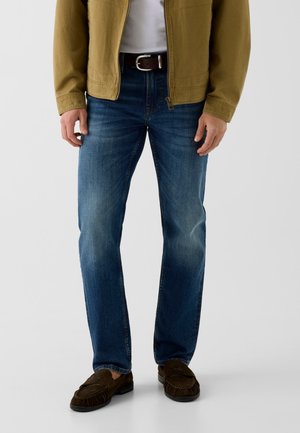 Homme portant un jean bleu, une ceinture marron, des mocassins marron, une chemise blanche et une veste marron clair, debout sur un fond clair uni.
