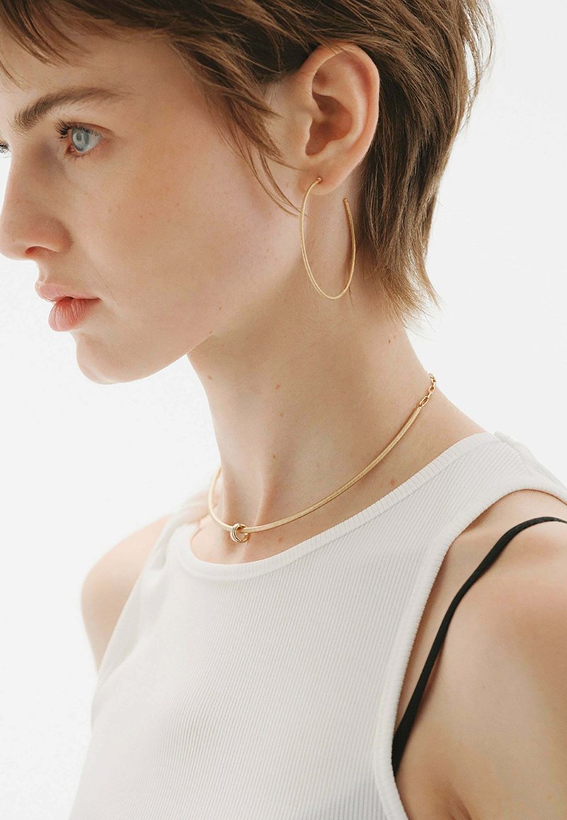 Pendientes de aro de oro y un collar choker de oro en una modelo. El choker presenta un acento circular. La modelo lleva una blusa blanca de canalé.