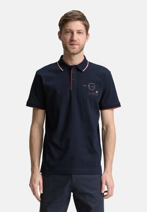 Man in een marineblauw poloshirt met korte mouwen, versierd met rode en witte accenten op de kraag en mouwen, staand tegen een witte achtergrond.
