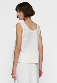 Donna con i capelli castani corti, che indossa un top bianco senza maniche e pantaloni bianchi, girata leggermente con un'espressione neutra.