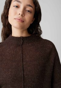 Brauner Strickpullover mit hohem Kragen, mit einer zentralen vorderen Naht und einer weichen, strukturierten Oberfläche. Keine sichtbaren Verschlüsse oder Muster.