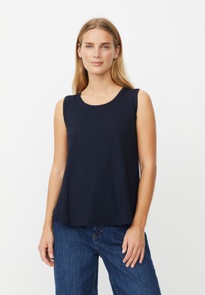 Giovane donna con lunghi capelli biondi che indossa una maglia senza maniche blu navy e jeans blu, in piedi davanti a uno sfondo bianco uniforme.