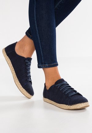 Persona che indossa jeans di denim blu scuro e scarpe in tela blu navy con suole in juta intrecciata su un pavimento bianco.