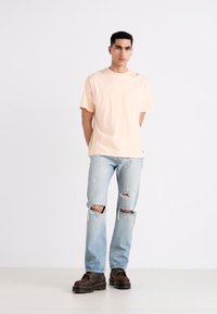 Světle broskvové tričko s kulatým výstřihem, světlé modré džíny s trhlinami na kolenou a tmavě hnědé boty. Jednoduchý design, volný střih.