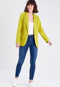 Blazer verde con un corte ajustado, diseño de un botón y dos bolsillos frontales, combinado con jeans ajustados azules y zapatillas blancas.