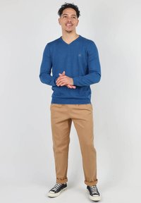 Pull bleu en V, pantalon beige, baskets montantes noires. Les textures semblent lisses, avec des détails de couture subtils sur le pull.