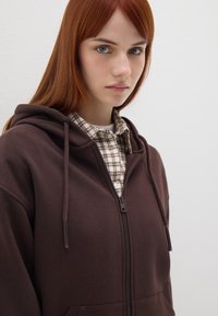 Sudadera marrón con cremallera y cordones, de tejido texturizado, con bolsillos delanteros. Superpuesta con una camisa de cuadros en tonos beige y marrón por debajo.