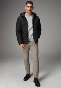 Sort puffjakke med hætte, prydet med et quiltet design. Bæres over en lys grå sweatshirt og kombineres med lyse beige bukser og sorte sko.