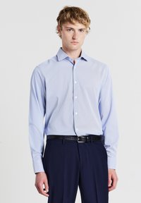 Giovane uomo con capelli biondi, che indossa una camicia da uomo azzurro chiaro con bottoni, una cintura nera e pantaloni blu scuro su uno sfondo neutro.