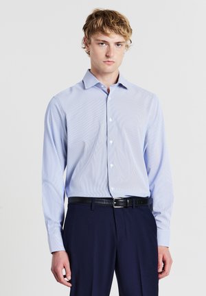Jeune homme aux cheveux blonds portant une chemise habillée bleu clair à boutons, une ceinture noire et un pantalon bleu marine foncé sur un fond uni.