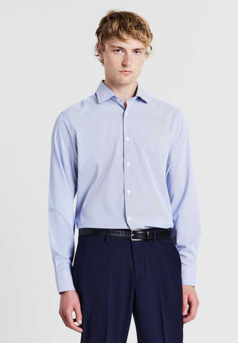 Giovane uomo con capelli biondi, che indossa una camicia da uomo azzurro chiaro con bottoni, una cintura nera e pantaloni blu scuro su uno sfondo neutro.