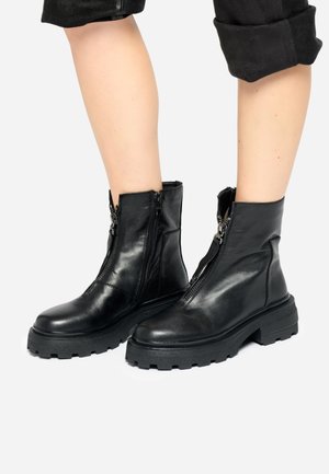 Personne portant des bottines en cuir noir avec des fermetures éclair frontales et des semelles en caoutchouc épaisses, associées à un pantalon noir retroussé.