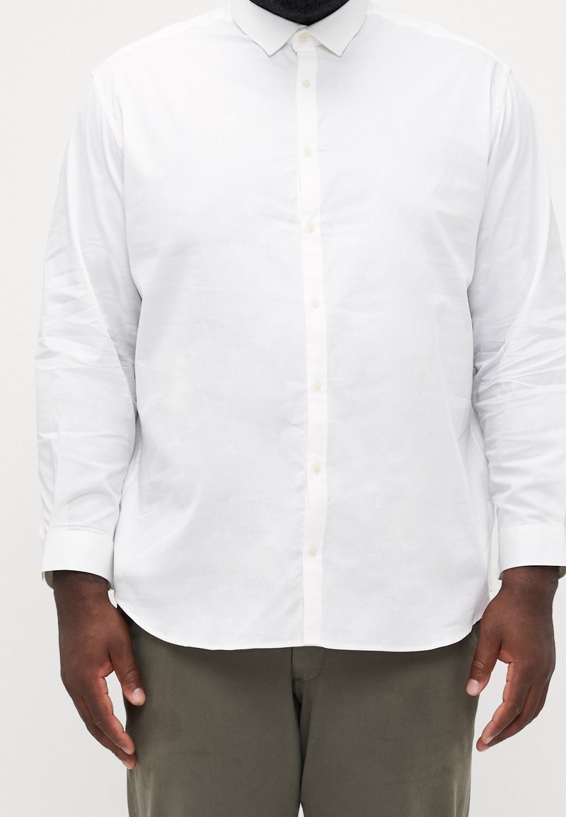 Homme portant une chemise blanche à manches longues boutonnée et un pantalon vert olive, debout devant un fond clair uni.