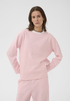 Giovane donna con capelli castani che indossa una felpa a maniche lunghe rosa chiaro e pantaloni abbinati, in piedi con le mani sui fianchi davanti a uno sfondo semplice.