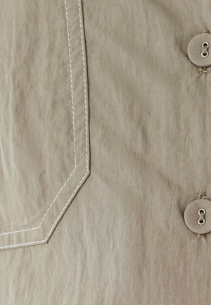 Tejido beige con una superficie texturizada; cuenta con costuras blancas y un botón redondo con centro hueco, acentuando un bolsillo de bordes afilados.