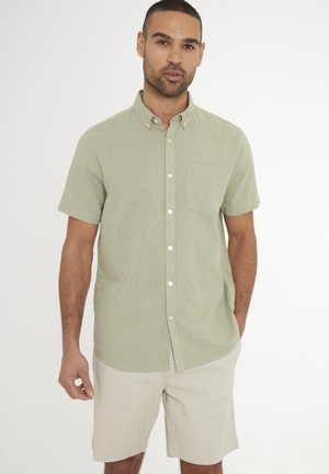 Mężczyzna w jasnozielonej koszuli na krótki rękaw z guzikami i beżowych krótkich spodenkach stojący na białym tle.