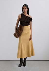 Femme portant un top brun foncé à une épaule, une jupe beige taille haute, des bottes noires, tenant un grand sac à main en daim brun contre un fond uni.