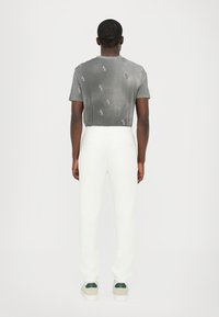 Grijze t-shirt met subtiel logo patroon, gecombineerd met witte broek. De outfit wordt compleet gemaakt met schoenen met groene accenten.
