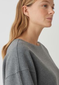 Grauer Wollpullover mit weitem Halsausschnitt, lockerer Passform und sichtbaren Nähten an den Ärmeln. Weiche Textur und minimalistisches Design.