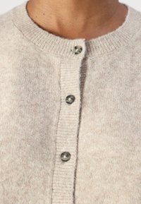 Lys beige cardigan laget av mykt strikkemateriale, med rund hals, frontlukking med knapper og strukturert ribbestrikk langs kragen.