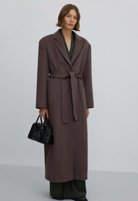 Lange bruine jas met een riem, grote revers en een gestructureerde afwerking. Draagt over een donkergroene outfit, aangevuld met een zwarte handtas.