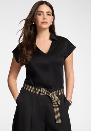 Frau mit langen braunen Haaren, die ein schwarzes kurzärmeliges Oberteil und schwarze Hose mit einem vorne gebundenen gestreiften Gürtel trägt, steht mit den Händen in den Taschen.