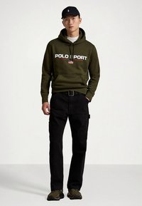 Πράσινο φούτερ με κουκούλα και το κείμενο "POLO SPORT", μαύρο παντελόνι cargo και χακί αθλητικά παπούτσια, σε συνδυασμό με μαύρο καπέλο, όλα σε casual εμφάνιση.