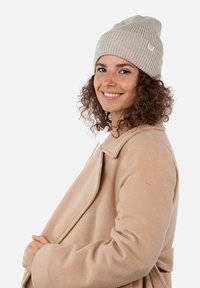 Gorro de punto acanalado beige con una pequeña etiqueta, llevado con un abrigo beige. El gorro tiene un diseño holgado y un patrón de punto texturizado.