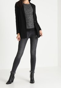 Černé na míru šité sako přes šedý pletený top, v kombinaci s tmavými úzkými džínami s třepeným lemem a černými kotníkovými botami.