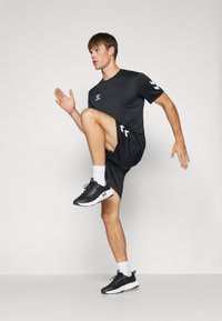 Camiseta atlética negra y pantalones cortos, con un logo y un diseño en chevron, combinados con zapatillas de correr negras y calcetines blancos, en una pose dinámica.