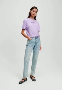 Camiseta corta de color púrpura claro con texto negro, combinada con jeans de tiro alto en azul claro y zapatos planos negros con lunares. Diseño sencillo y moderno.