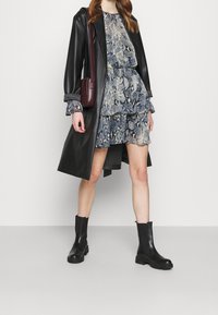 Černý kožený trenčkot přes modro-šedě vzorované šaty s volány, doplněný černými kotníkovými botami a burgundskou taškou přes rameno.