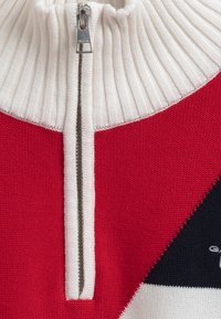 Maglione lavorato a maglia rosso e bianco con colletto alto a costine e chiusura a mezza zip; presenta motivi geometrici in contrasto tra il nero e il bianco.