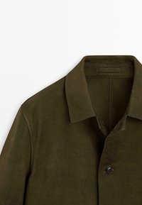 Olivenfarbene Wildlederjacke mit spitzem Kragen, frontaler Knopfverschluss und glatter Textur. Enthält ein Etikett am Kragen.